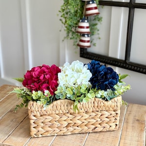 Könnte beinhalten: Ein rechteckiger Weidenkorb gefüllt mit leuchtenden Kunstblumen in Rot, Weiß und Blau, akzentuiert mit grünem Laub. Die Anordnung steht auf einer Holzoberfläche und erinnert an ein patriotisches Thema. Dekorative Glocken mit rot-weiß-blauen Streifen hängen im Hintergrund.