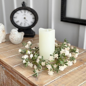 Puede incluir: Una composición decorativa sobre una mesa de madera. Presenta una vela color crema rodeada por una corona floral. Dos pequeñas figuras de pájaros y un reloj negro completan la escena, creando un ambiente rústico.
