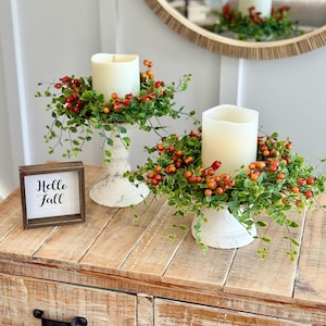 May include: Two white pillar candles on distressed white pedestal candle holders, each surrounded by a wreath of green leaves and orange berries. A small square wooden sign with the black text Hello Fall sits next to the candle holders.