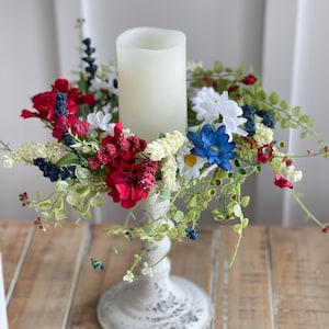 May include: A white candle holder with a white pillar candle surrounded by a red, white, and blue floral arrangement. The arrangement includes red geraniums, white daisies, and blue flowers.