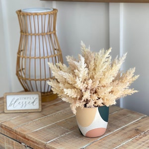 May include: A dried pampas grass bouquet in a cream, pink, green, and white geometric patterned ceramic planter. The planter is sitting on a wooden table with a sign that reads "simply blessed".
