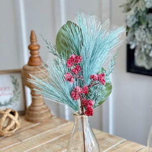 May include: A frosted blue pine sprig with red berries in a clear glass bottle. The bottle is sitting on a wooden table.