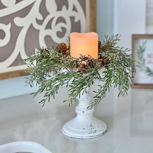 May include: A white pillar candle holder with a frosted glass candle and a green garland with faux snow. The garland is decorated with pine cones. The candle holder is on a white surface with a wooden sign that says "Merry Christmas" in the background.