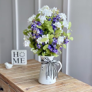 May include: A white pitcher with black and white striped ribbon tied around the handle, filled with a bouquet of white and purple flowers. The pitcher is sitting on a wooden table with a sign that says "HOME" on it.