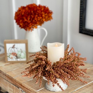 May include: A cream-colored pillar candle sits atop a distressed white pedestal surrounded by dried orange-brown foliage. A small framed picture of pumpkins and a white pitcher with orange flowers are in the background. The items are on a wooden table.