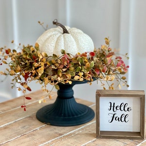 Könnte beinhalten: Weißer Kürbis mit einem Herbstlaubkranz auf einem schwarzen Sockel mit einem Holzschild mit der Aufschrift "Hello Fall" auf einem hellen Holztisch.