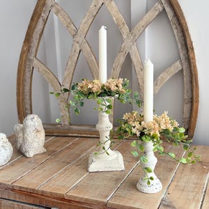 May include: Two off-white candle holders with cream-colored candles, adorned with faux greenery and flowers. The candle holders sit on a rustic wooden surface, with decorative bird figurines and a wooden window frame in the background.