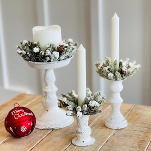 Op de afbeelding: Drie witte kandelaars met gematteerde glazen kaarsen en een besneeuwde groenblijvende krans. De kandelaars staan op een houten tafel met een rode kerstbel met de tekst "Merry Christmas".