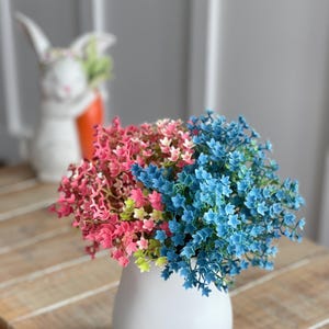 May include: A white ceramic vase with a bouquet of pink, green, and blue artificial flowers. The flowers are arranged in a cluster and are in a vase on a wooden surface.