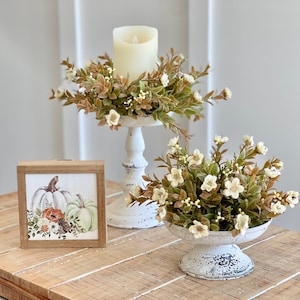 May include: Two white metal candle holders with a distressed finish. The larger holder has a pillar candle and a wreath of green leaves and white flowers. The smaller holder has a cluster of white flowers and green leaves. A framed print of white pumpkins and flowers sits on a wooden table.