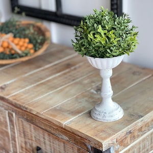 May include: A white pedestal with a green topiary plant on top. The pedestal is on a wooden table with drawers.