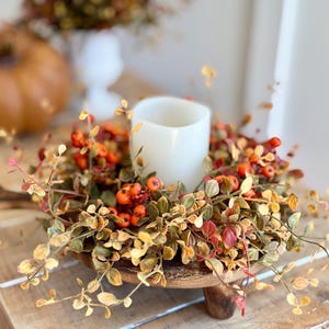 May include: A wooden pedestal with a rustic finish holds a fall-themed wreath. The wreath is made of faux greenery with small orange berries and a white pillar candle in the center.