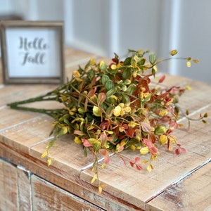 Puede incluir: Un ramo de vegetación artificial con hojas verdes, amarillas y rojas. La vegetación está sobre una superficie de madera clara con un letrero que dice "Hello Fall".