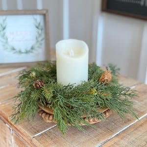 May include: A cream-coloured LED candle sits on a wooden beaded base, encircled by a green and brown evergreen wreath with pine cones. A framed sign with "Merry Christmas" is in the background. The arrangement is on a wooden surface.