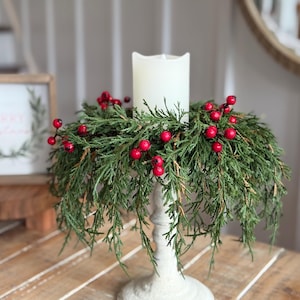 May include: A white pillar candle sits in a tall, off-white candlestick holder, surrounded by a wreath of green cedar branches and bright red berries. The candle features a wavy top edge, and the greenery drapes downward around the base.