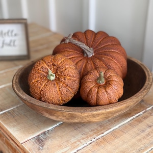 May include: Three textured, orange-brown decorative pumpkins of varying sizes nestled inside a round, dark brown wooden bowl. A small, square wooden sign with the text "Hello Fall" in black script is positioned in the background.