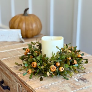 May include: A decorative fall-themed arrangement featuring a cream-colored candle surrounded by a green and orange berry wreath. A small pumpkin and a white rectangular object are in the background, all set on a rustic wooden table.