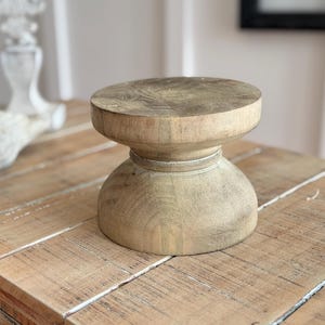 May include: A light-colored, wooden pedestal with a round top and a bulbous base. The pedestal is made of natural wood and sits on a wooden table. A decorative bird figurine is in the background.