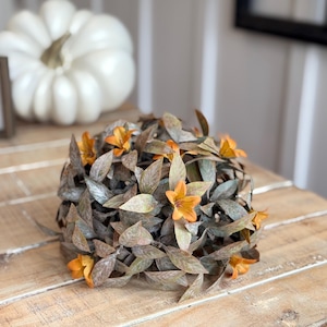 Arranjo de outono com folhagem rústica e flores laranja/mostarda, ideal para preencher vasos e decorar ambientes outonais. Formato de meia esfera.