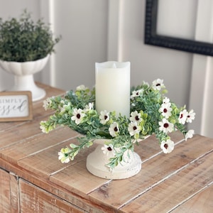 Könnte beinhalten: Weiße Kerze in Säulenform mit einem weißen Blumenkranz auf einem weißen Sockel. Die Kerze und der Kranz stehen auf einem Holztisch mit einem Schild, auf dem "Simply Blessed" steht.