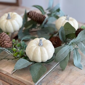 May include: A white pumpkin wreath with green leaves and brown pine cones. The wreath is made of artificial materials and is perfect for fall decor.