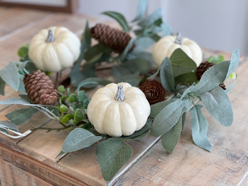 May include: A white pumpkin wreath with green leaves and brown pine cones. The wreath is made of artificial materials and is perfect for fall decor.