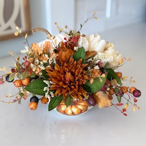 May include: A decorative floral arrangement featuring a central rust-coloured dahlia, cream-coloured flowers, and small orange and dark purple berries. The arrangement is set within a small, round ceramic pumpkin-shaped container with a stem.