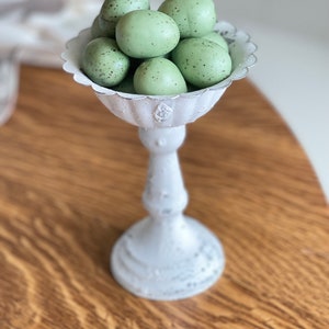 May include: A white ceramic pedestal with a scalloped edge bowl filled with speckled green Easter eggs.