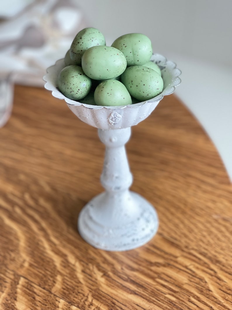 May include: A white ceramic pedestal with a scalloped edge bowl filled with speckled green Easter eggs.