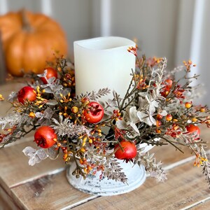 May include: A white pillar candle surrounded by a fall-themed floral arrangement. The arrangement includes red berries, orange and yellow accents, and faux foliage. The candle sits on a distressed white metal base. An orange pumpkin is in the background.