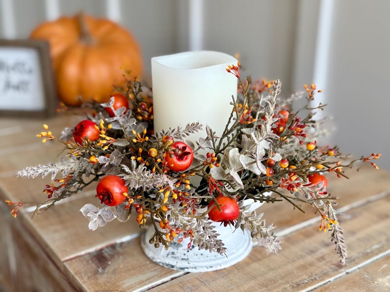 May include: A white pillar candle surrounded by a fall-themed floral arrangement. The arrangement includes red berries, orange and yellow accents, and faux foliage. The candle sits on a distressed white metal base. An orange pumpkin is in the background.