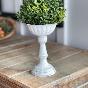 May include: A white ceramic pedestal with a fluted bowl top, holding a green artificial topiary plant.