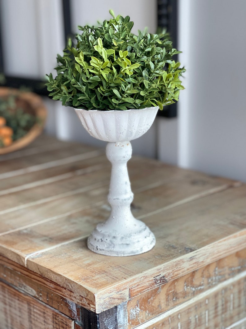 May include: A white ceramic pedestal with a fluted bowl top, holding a green artificial topiary plant.