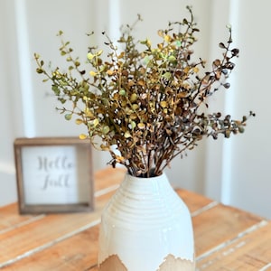 May include: A white and tan ceramic vase with a textured design, holding a bundle of dried greenery. The vase sits on a wooden surface. A small wooden framed sign with the words "Hello Fall" is in the background.