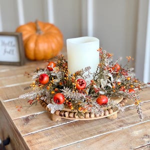 May include: A fall-themed centerpiece featuring a white candle surrounded by faux foliage, berries, and small red apples. The arrangement sits on a wooden tray with a beaded edge. An orange pumpkin and a framed sign that says "Hello Fall" are in the background.