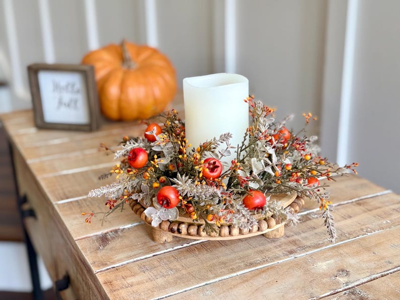 May include: A fall-themed centerpiece featuring a white candle surrounded by faux foliage, berries, and small red apples. The arrangement sits on a wooden tray with a beaded edge. An orange pumpkin and a framed sign that says "Hello Fall" are in the background.
