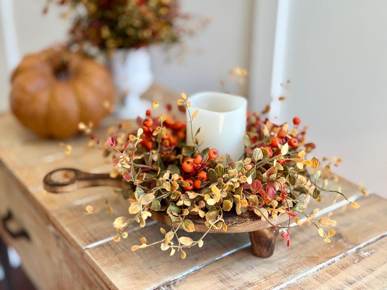 May include: A wooden pedestal with a fall-themed wreath featuring orange berries and gold leaves. A white pillar candle sits in the center of the wreath.