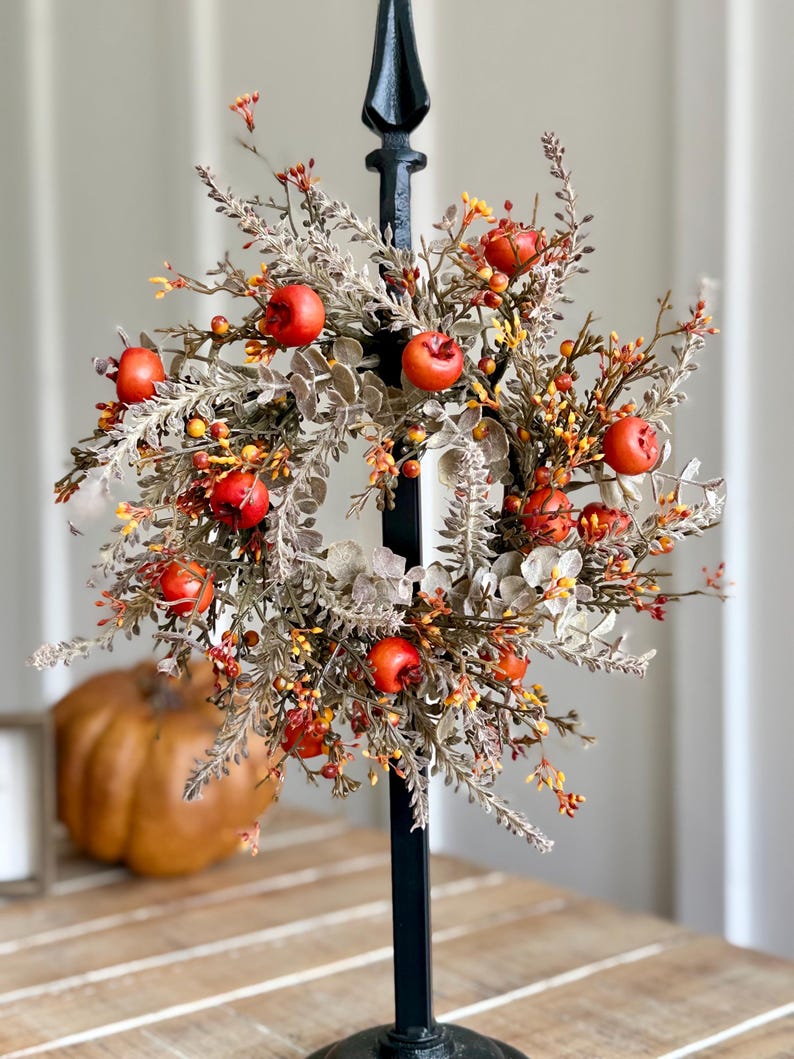 May include: A fall-themed wreath with faux red apples, orange berries, and muted green foliage. The wreath is mounted on a black metal stand, perfect for seasonal home decor. The wreath is a circular shape.