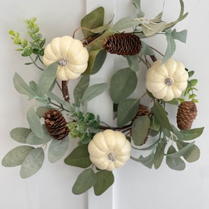 May include: A white faux pumpkin wreath with green leaves and brown pine cones.