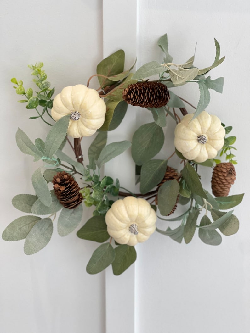 May include: A white faux pumpkin wreath with green leaves and brown pine cones.