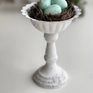 May include: A white ceramic pedestal with a bird's nest and three light blue speckled eggs. The pedestal has a fluted design and a round base.