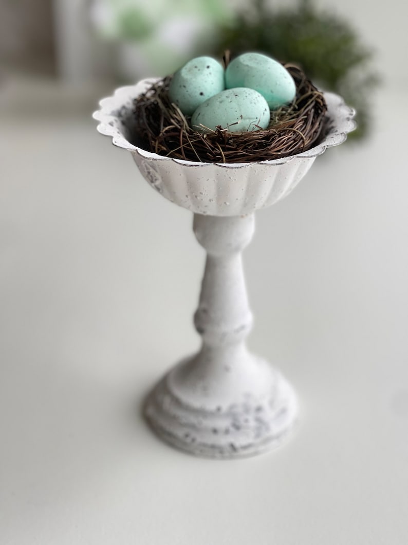 May include: A white ceramic pedestal with a bird's nest and three light blue speckled eggs. The pedestal has a fluted design and a round base.