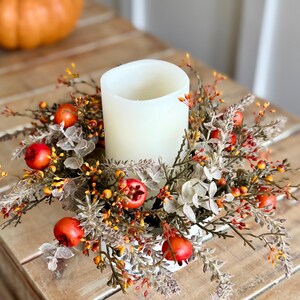 May include: A white pillar candle centerpiece surrounded by a wreath of fall foliage, including red berries, orange and yellow accents, and gray-green leaves. The candle sits on a white base, creating a warm, autumnal display.