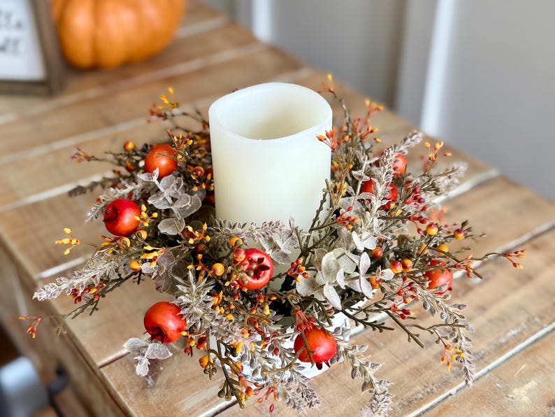 May include: A white pillar candle centerpiece surrounded by a wreath of fall foliage, including red berries, orange and yellow accents, and gray-green leaves. The candle sits on a white base, creating a warm, autumnal display.