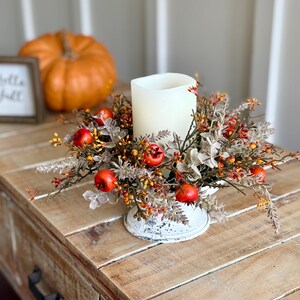 May include: A fall-themed centerpiece with a large white candle surrounded by faux berries and foliage. The arrangement sits in a distressed white metal base on a wooden table. An orange pumpkin and a framed sign that says "Hello Fall" are in the background.