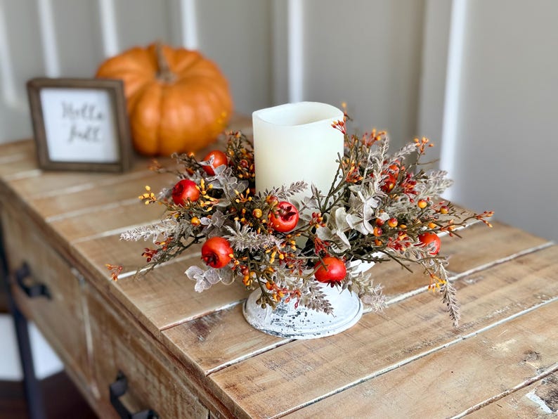 May include: A fall-themed centerpiece with a large white candle surrounded by faux berries and foliage. The arrangement sits in a distressed white metal base on a wooden table. An orange pumpkin and a framed sign that says "Hello Fall" are in the background.