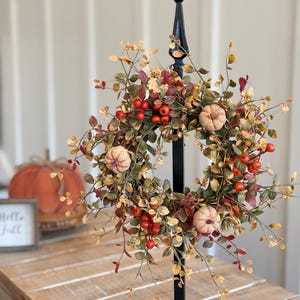 May include: A seasonal wreath featuring artificial pumpkins, red berries, and green and yellow foliage. The wreath is displayed on a black metal stand. A small sign with the words "Hello Fall" is in the background.