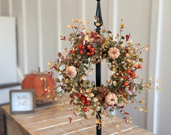 May include: A fall-themed wreath with faux pumpkins, red berries, and green and yellow leaves. The wreath is mounted on a black metal stand. A small sign that says "Hello Fall" is visible in the background.