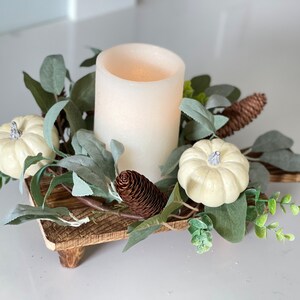 May include: A white pillar candle sits in the center of a wooden tray surrounded by faux greenery, white pumpkins, and pine cones.