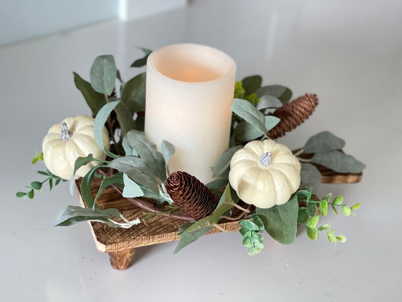 May include: A white pillar candle sits in the center of a wooden tray surrounded by faux greenery, white pumpkins, and pine cones.